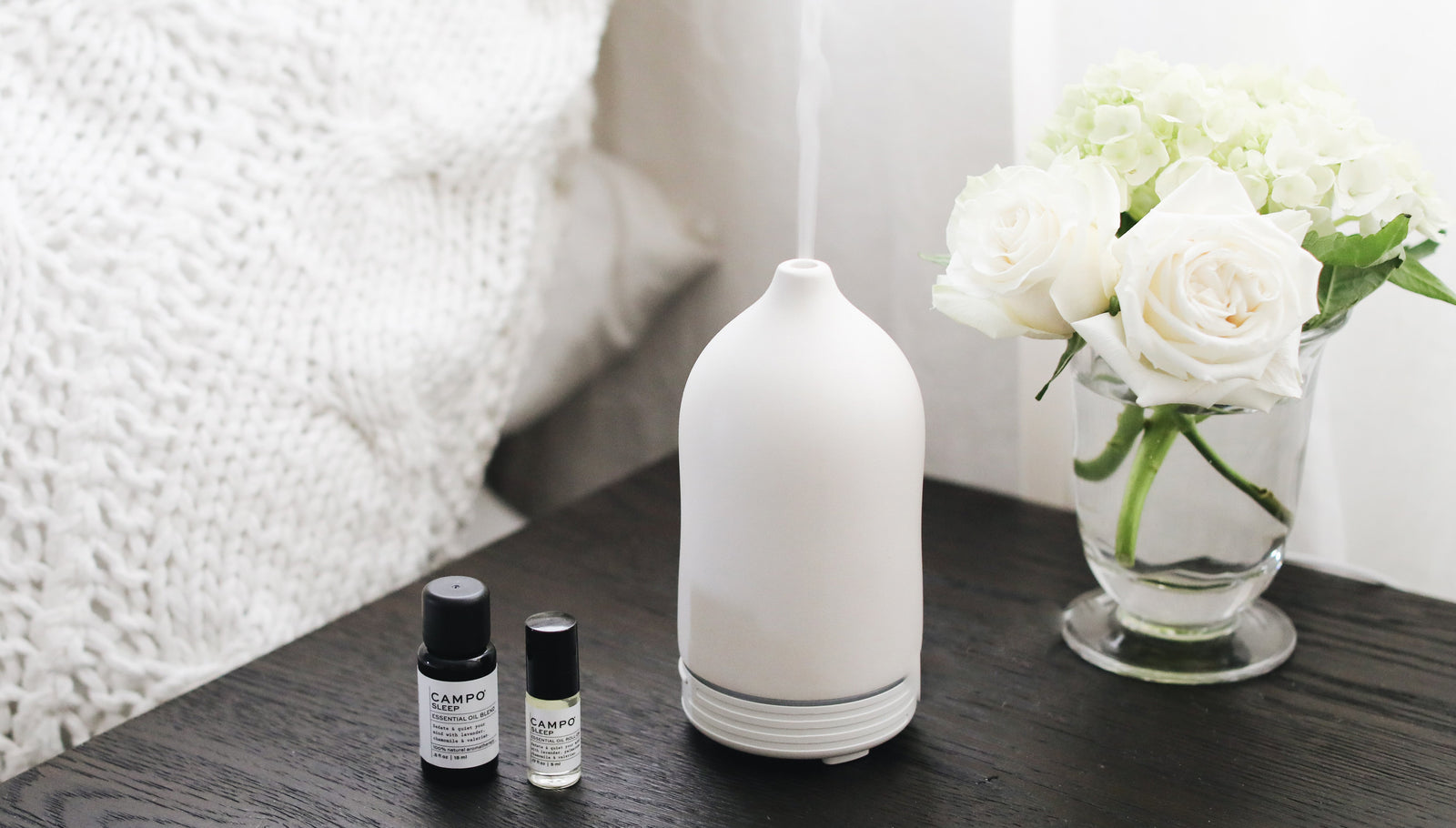 White diffuser and small bottles on a wooden surface with white flowers in the background